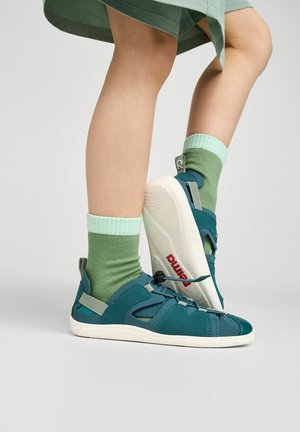Dziecko noszące turkusowe sandały z regulowanymi paskami i zielone skarpetki, stojące na jednej nodze z uniesioną stopą, pokazującą białą podeszwę z czerwonym logo.