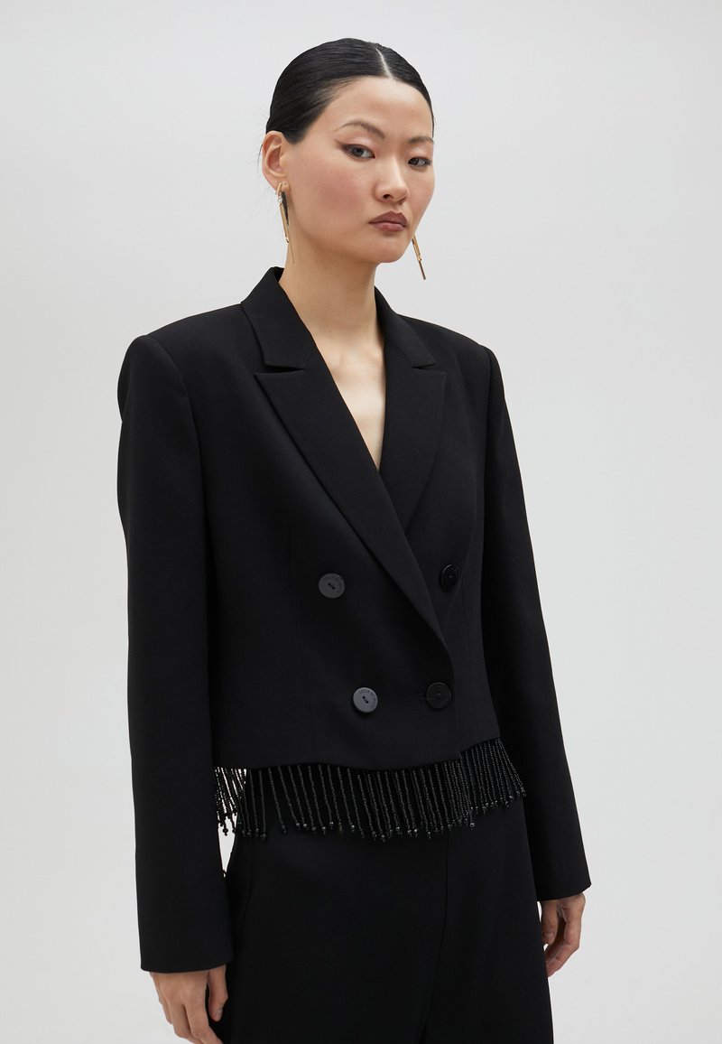 Svart kort dubbelknäppt blazer med två stora knappar och en fransförsedda, pärlprydd nederkant. Slät tyg med skarpa slag och långa ärmar.