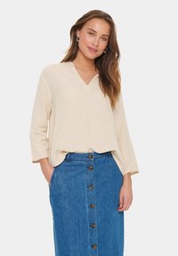 Blusa beige con scollo a v e maniche a tre quarti, abbinata a una gonna di jeans a vita alta con dettagli di bottoni Frontali.