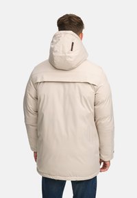 Veste matelassée beige avec capuche, dotée d'un empiècement dorsal, d'une texture lisse et de ferrures ajustables. Manches longues et coupe mi-longue.