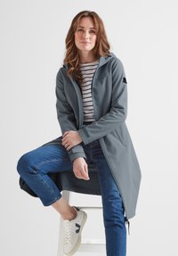 Long, gray softshell jacket with a hood, zip closure, and side pockets, paired with a striped top, blue jeans, and white sneakers.