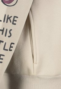 Beige Sweatshirt mit einer Seiten Tasche und teilweise sichtbarem schwarzen Text, der "LIKE THIS LITTLE" auf dem Ärmel liest.