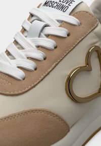 Κρεμ και μπεζ αθλητικό παπούτσι με λευκά κορδόνια, χρυσό έμβλημα καρδιάς στο πλάι και την ένδειξη "Love Moschino" στη γλώσσα.