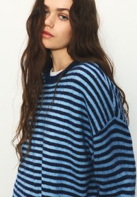 Pruhovaný sveter zo soft tkaniny; obsahuje striedavé pásy svetlomodrej a námorníckej modrej farby, s voľným strihom a okrúhlym výstrihom.
