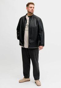 Sort læderjakke med lynlåslukning, kombineret med en lysegrå t-shirt med grafik og mørkegrå bukser. Brune ruskindssko, stående mod en hvid baggrund.