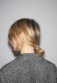 Vævet sort og hvidt hørfiskebensfrakke, set bagfra. Håret stylet i en lav hestehale, sikret med et hårbånd. Ensfarvet grå baggrund.