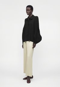Mujer con una blusa negra suelta de manga larga con mangas abullonadas, pantalones anchos color crema y zapatos planos marrón oscuro, de pie sobre un fondo blanco.