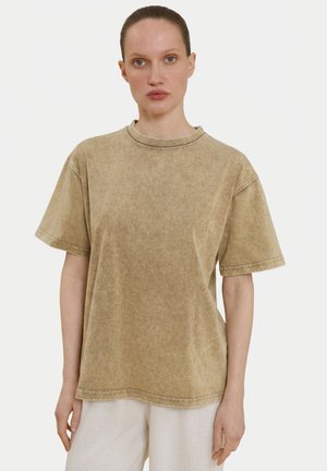 Femme aux cheveux tirés en arrière portant un t-shirt beige clair à manches courtes, ample et délavé, ainsi qu’un pantalon clair, debout devant un fond uni.