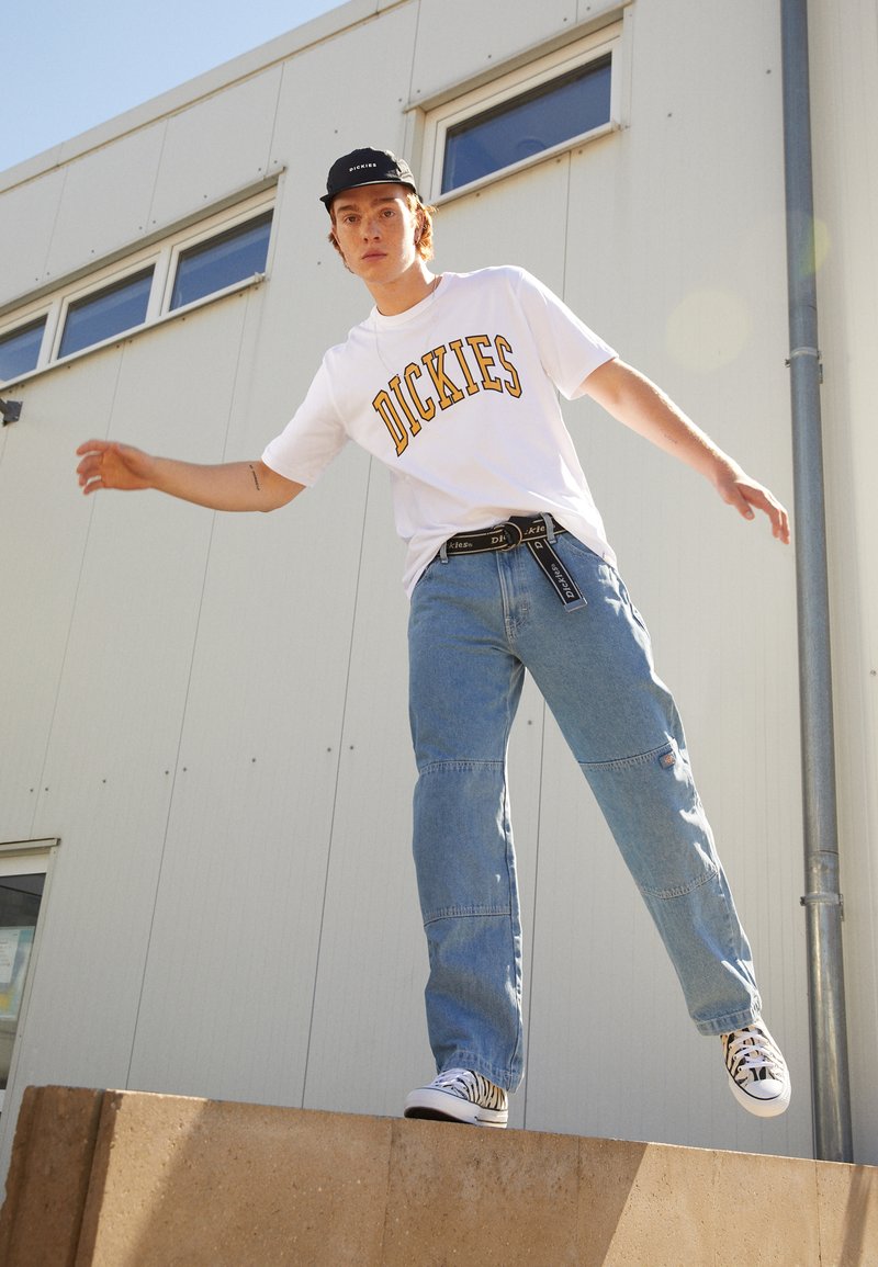 Hvit T-skjorte med gult "DICKIES"-logo, lyseblå baggy jeans med lapp, svart caps, og svart-hvite mønstrede sneakers, stående på en kant.