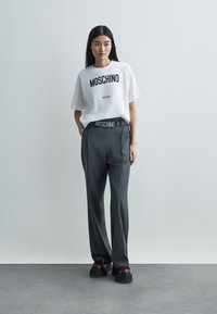 Λευκό μπλουζάκι με μαύρο κείμενο "Moschino Milano", σε συνδυασμό με γκρι παντελόνι που διαθέτει ζώνη "Moschino", και χοντρά μπορντό πλατφόρμες.