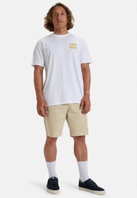 Hvid bomuldst-shirt med korte ærmer og lille gul logo på brystet, kombineret med lyse beige fløjlsbukser og marineblå sneakers.