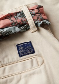 Beige stof met een blauw "Superdry MFG Co" label. Accenten van roze en turquoise gestreept stof op de band met een zachte textuur.