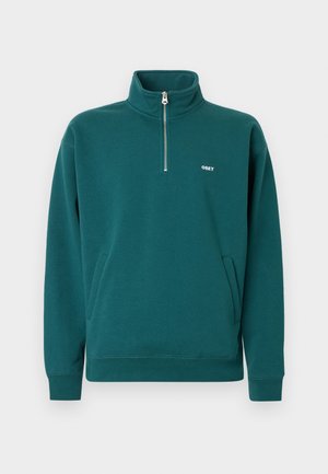 Felpa teal con zip completa, collo alto, tasche frontali e logo bianco "OBEY" sul petto. Tessuto morbido con finitura texturizzata.