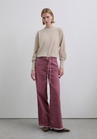 Pull en maille beige à manches longues, associé à un jean large rose avec un motif zèbre et un ourlet effiloché. Chaussures marron pointues.