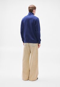 Μπλε μπλούζα με ψηλό γιακά, συνδυασμένη με μπεζ παντελόνι φαρδιάς ποδός, με λεία υφή και εμφανείς ραφές στις τσέπες.