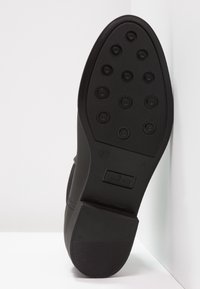 Suela de zapato negra con una superficie texturizada, que presenta puntos de tracción circulares y un tacón plano. Marcada con el logo de la marca y detalles de tamaño.