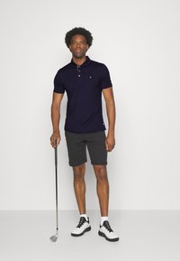 Mornarska polo majica z diskretnim logotipom, v kombinaciji s temnosivimi kratkimi hlačami in belimi golf čevlji. Model drži golf palico, stoji na svetli podlagi.