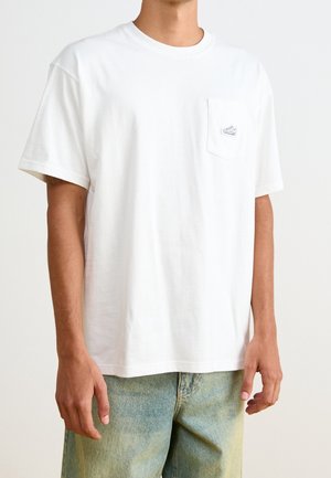 Personne portant un T-shirt blanc uni à manches courtes avec un petit logo de basket sur la poche poitrine et un jean en denim bleu clair.