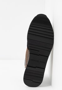 Semelle d'une chaussure noire avec une épaisse semelle crantée et le nom de la marque Michael Kors embossé sur la semelle, inclinée contre un mur blanc.