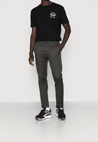 T-shirt nera a maniche corte con grafica bianca, pantaloni grigi slim-fit e sneaker nere con dettagli grigi e bianchi, in piedi su una superficie chiara.