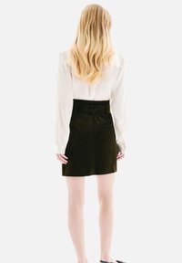 Femme avec de longs cheveux blonds portant un chemisier blanc à manches longues rentré dans une jupe noire taille haute, se tenant dos.