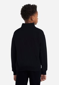 Gutt med kort krøllete hår som har på seg en svart langermet genser, står med ryggen til mot en lys hvit bakgrunn.