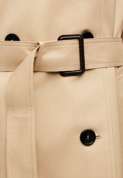 Μπεζ παλτό τσέντρα με ταιριαστή ζώνη που περνάει μέσα από μια σκοτεινή ορθογώνια αγκράφα και μεγάλα μαύρα κουμπιά μπροστά.