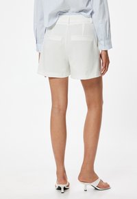 Hvide skræddersyede shorts med elastisk talje, flade baglommer og en glat tekstur. Bæres med hvide åben-toe hæle.