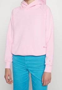 En person bär en rosa huvtröja och blå manchesterbyxor, stående mot en neutral bakgrund och visar en avslappnad outfit.