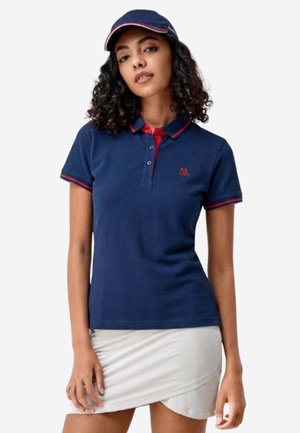 Frau trägt ein marineblaues Polo-Shirt mit rotem Besatz und passender Kappe, kombiniert mit einem weißen Wickelrock, stehend vor einem schlichten Hintergrund.