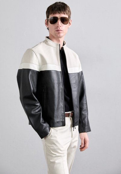 Black and cream leather jacket with color-blocked design, high collar, front zipper, and side pockets. Model wears beige pants and sunglasses.