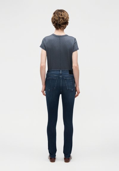 Άτομο με κοντά μαλλιά στέκεται γυρισμένο πλάτη, φορόντας γκρι κοντομάνικη μπλούζα, σκούρο μπλε εφαρμοστά τζιν και καφέ παπούτσια πάνω σε απλό φόντο.