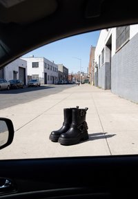 Paar schwarze Lederstiefel mit Schnallen auf dem Bürgersteig neben einer leeren Straße, gesehen von innen eines Autos.