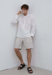 Biała lniana koszula z długim rękawem i zapięciem na guziki, w połączeniu z jasnobeżowymi szortami i czarnymi sandałami, na tle gładkiej ściany.