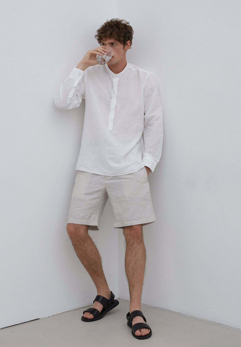 Biała lniana koszula z długim rękawem i zapięciem na guziki, w połączeniu z jasnobeżowymi szortami i czarnymi sandałami, na tle gładkiej ściany.