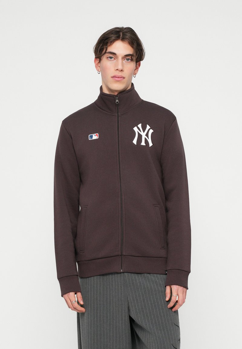 Temno rjava jakna z zadrgo in visokim ovratnikom, ki vsebuje belo vezeno oznako New York Yankees in oznako MLB. Ima stranske žepe in rebraste manšete.