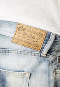 Džínové kalhoty s světlým modrým, opotřebovaným vzhledem. Obsahují hnědou koženou visačku s nápisem "Polo Ralph Lauren" s embosovanými detaily a švy.