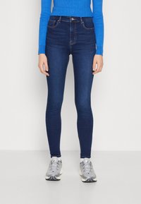 Eng skinny Jeans aus dunkelblauem Denim mit hoher Taille, Fronttaschen und dezenten Nähten. Kombiniert mit grauen Sportschuhen.