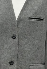 Veste grise en mélange de laine avec une finition texturée, présentant deux boutons noirs et une poche poitrine unique avec une petite ouverture.