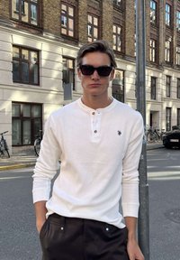 Mand iført hvid henley-bluse med lange ærmer og sorte solbriller, stående på en bygade med murstensbygninger og cykler i baggrunden.