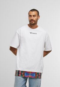 Bărbat cu păr scurt și barbă, purtând un tricou alb Colucci suprapus peste o cămașă lungă cu un model colorat și blugi de culoare albastru deschis.