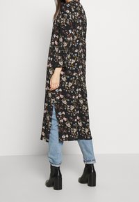 Kvinna som står i ljusblå jeans, svart blommig lång tunika med sidospalt och svarta klackstövlar mot en enfärgad vit bakgrund.