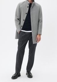 Cappotto di lana grigio con colletto alla coreana, chiusura con bottoni e tasche laterali; indossato sopra un maglione blu navy e abbinato a pantaloni scuri a quadri e scarpe nere.