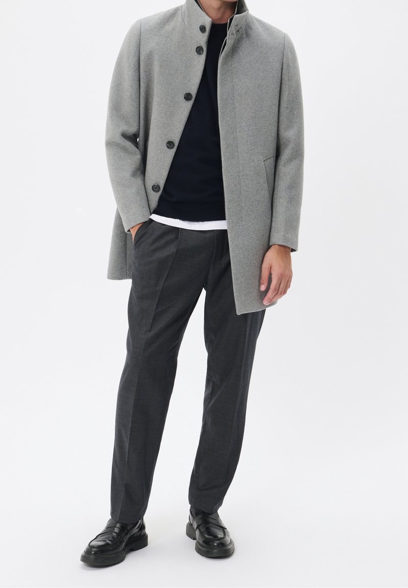 Cappotto di lana grigio con colletto alla coreana, chiusura con bottoni e tasche laterali; indossato sopra un maglione blu navy e abbinato a pantaloni scuri a quadri e scarpe nere.