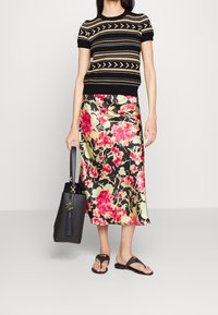 Svart kortärmad tröja med geometriska mönster, tillsammans med en blommig satinskjol i rosa och gröna nyanser. En svart tote-väska och flip-flop-sandaler fulländar looken.