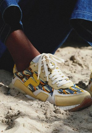 Personne portant des baskets à motifs colorés avec des accents jaunes et orange, assise sur un sol sablonneux, habillée d'un jean bleu retroussé.