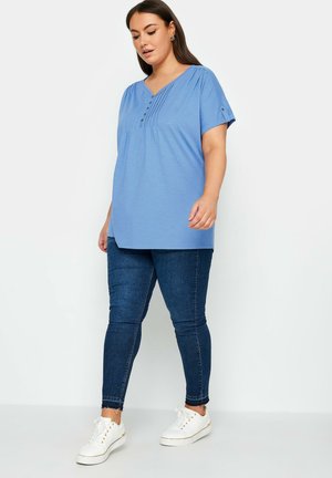 Blauw T-shirt met korte mouwen, een V-hals en knoopdetail, met verticale plooien. Gecombineerd met donkerdenim skinny jeans en witte sneakers.