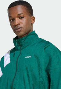 Grön lättviktsjacka med vita detaljer, hög krage och dragkedjedetalj. Har Adidas-logotypen på bröstet. Texturerat tyg.