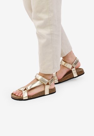 Sandales en métal doré avec des brides réglables, semelle intérieure en liège et semelle en caoutchouc noir, présentant un design ouvert à l'avant et à l'arrière.