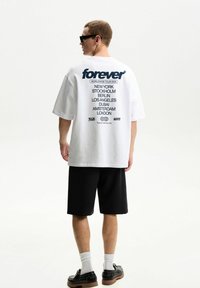 White oversized t-shirt featuring black "forever" text and city names printed on the back. Paired with black shorts and black shoes.